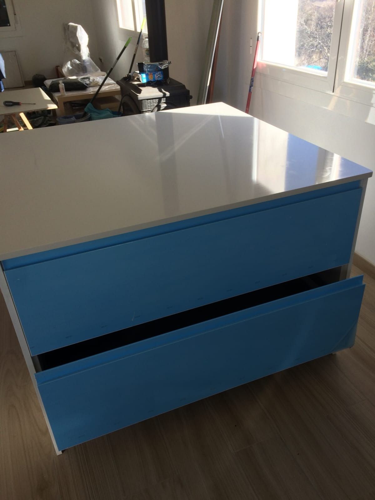 En una habitación, sobre un suelo de madera, hay un armario azul brillante de dos cajones con una encimera blanca.