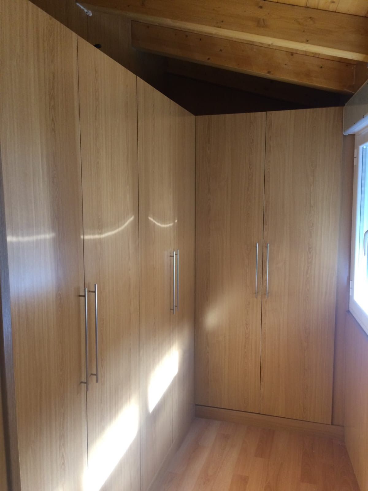 Un rincón de una habitación con armarios altos de madera clara con tiradores verticales plateados y suelo de madera noble.