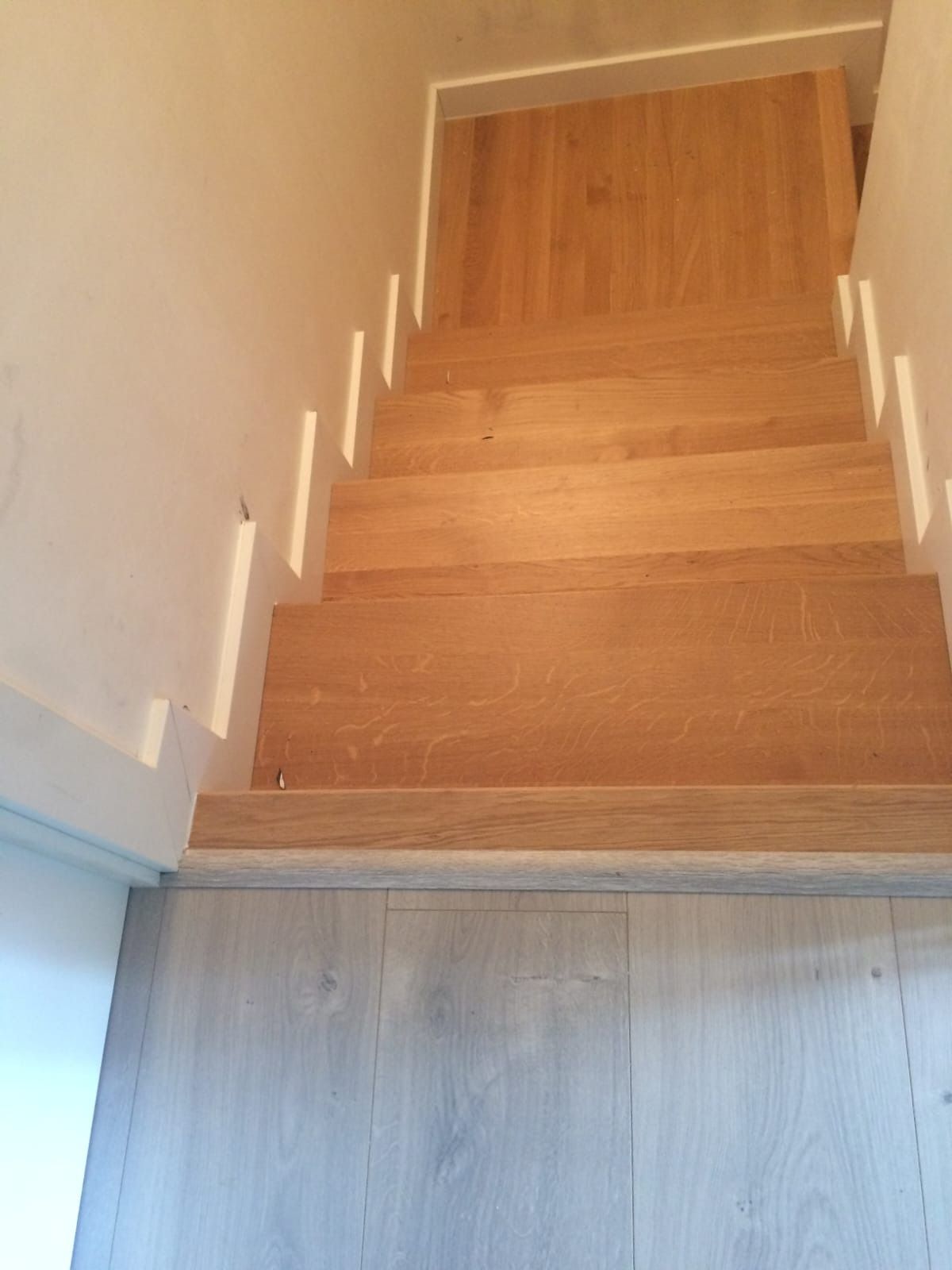 Escalera de madera que baja desde un rellano hasta un pasillo con suelo de madera clara y paredes blancas.