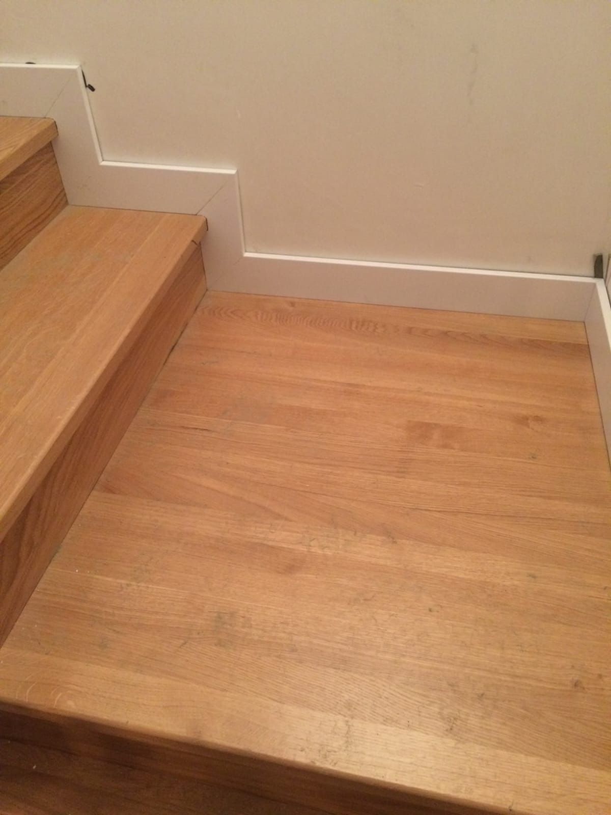 Una escalera de madera de roble claro conduce a un piso con zócalos blancos contra una pared lisa.