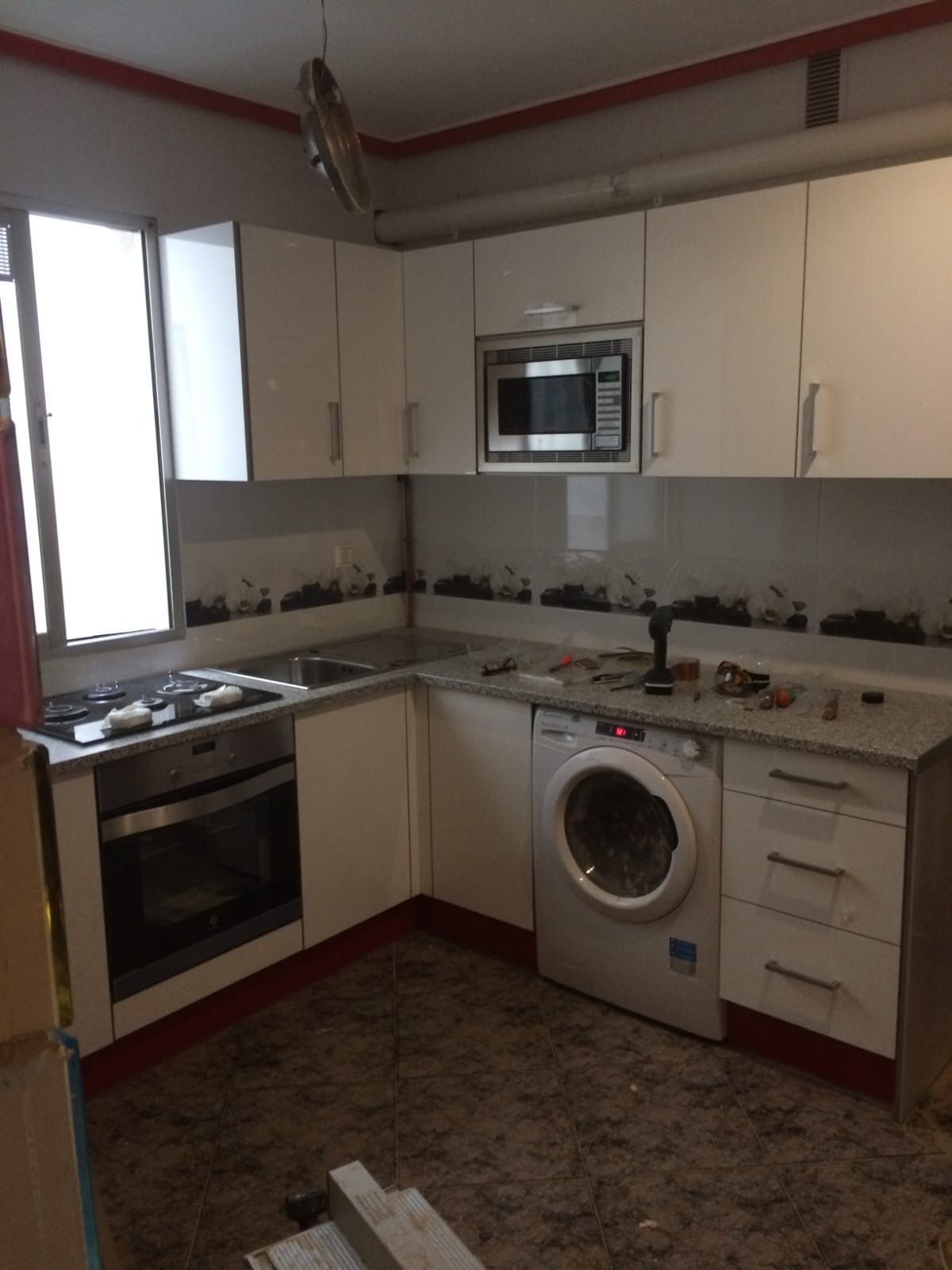 Una cocina con armarios blancos, horno empotrado, microondas y lavadora, con un salpicadero estampado.