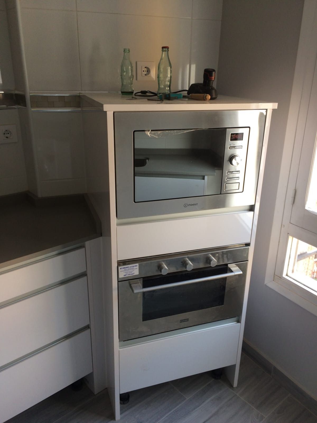 Un microondas y horno empotrados de acero inoxidable en un mueble de cocina blanco junto a una ventana.