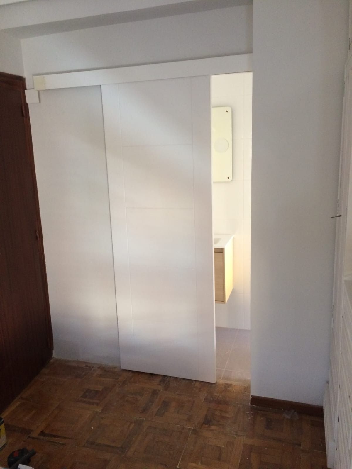 Una puerta corredera blanca, entreabierta, deja ver un baño con lavabo y espejo, sobre un suelo de parqué.