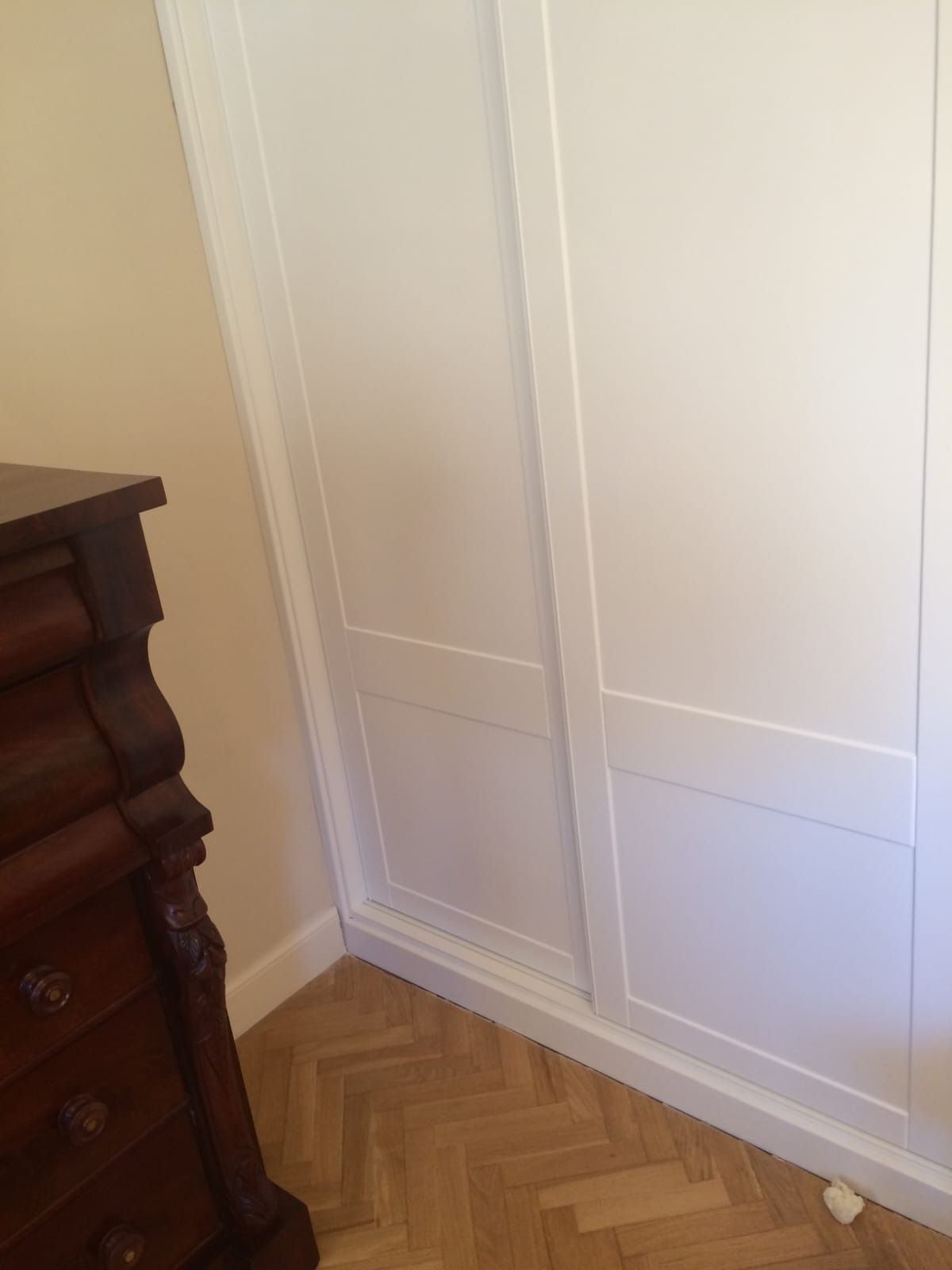 Una cómoda de madera junto a la puerta de un armario blanco sobre un suelo de madera con diseño de espiga.