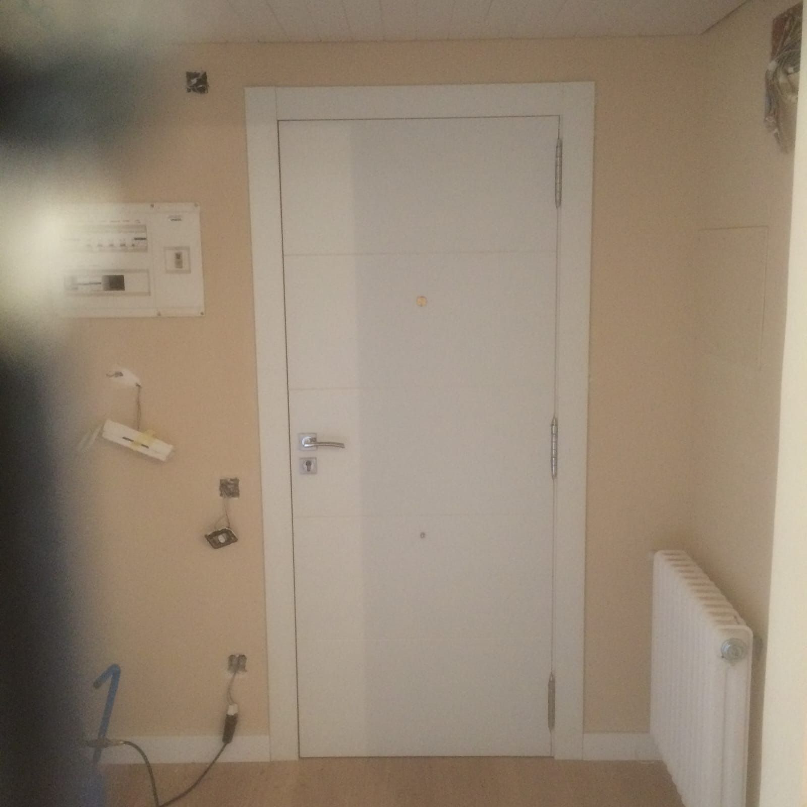 Una puerta blanca en un pasillo beige con un panel eléctrico montado en la pared, un radiador y varios enchufes.