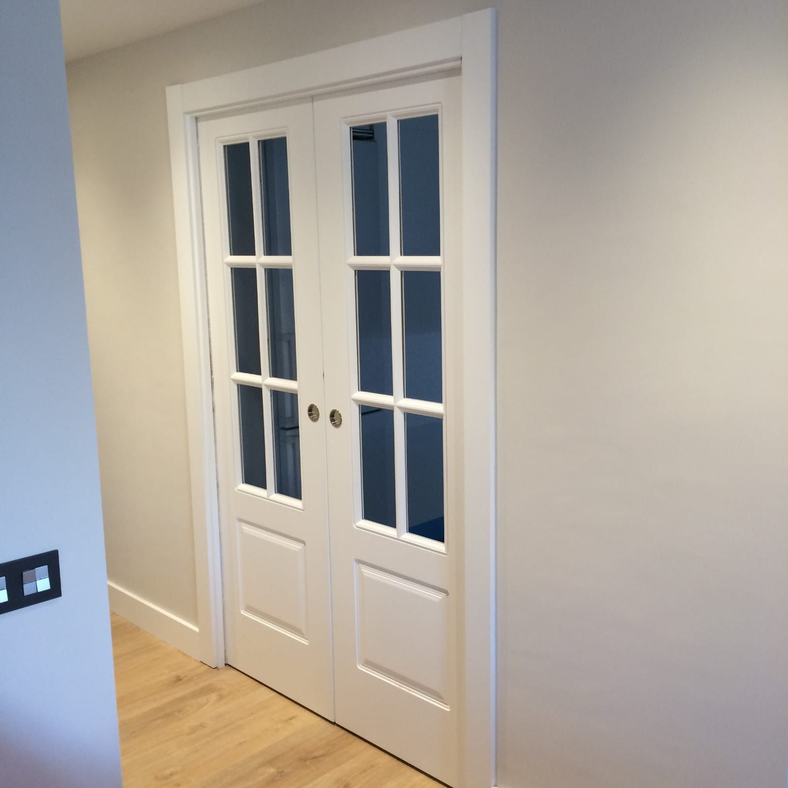 Puertas dobles francesas blancas con paneles de vidrio de seis hojas enmarcadas en blanco sobre una pared gris claro.