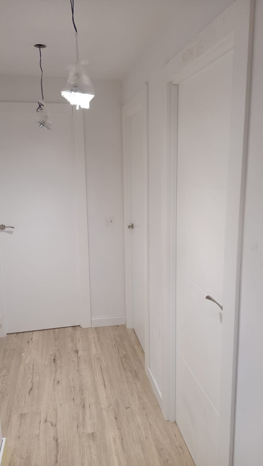 Un pasillo luminoso con puertas blancas, suelo de madera clara y dos lámparas colgantes que cuelgan del techo.
