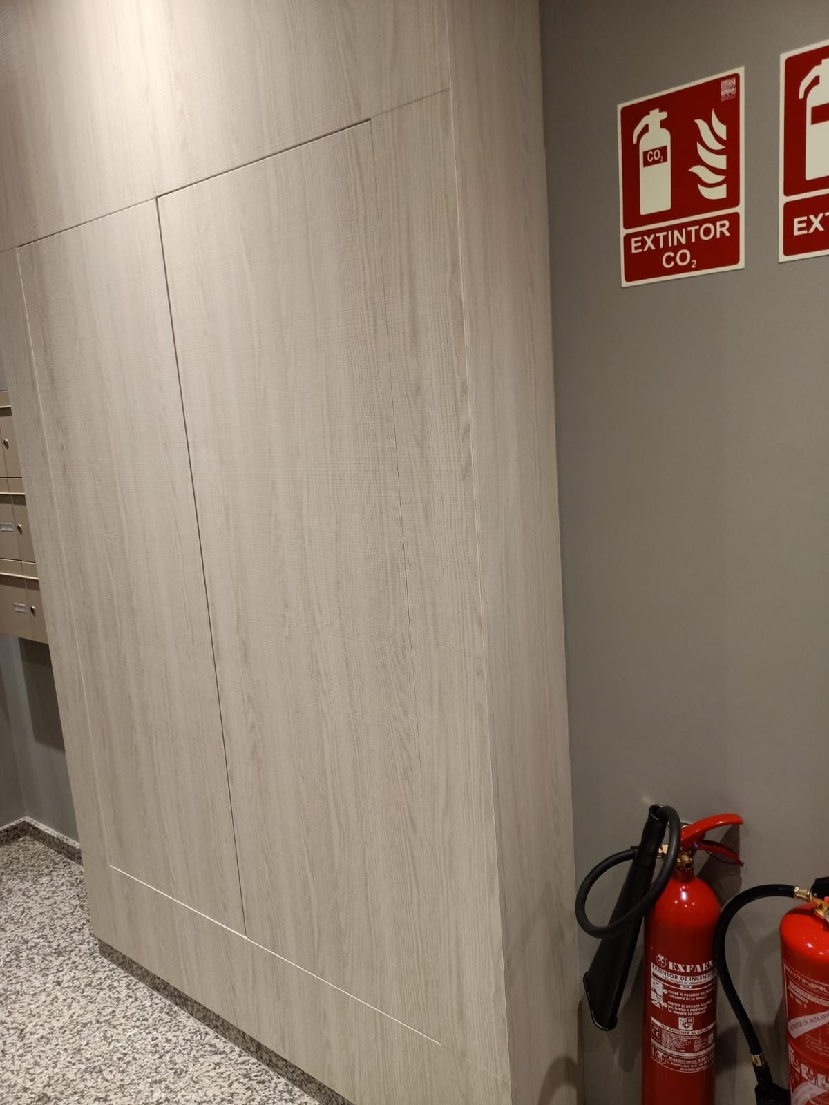 Un armario de madera clara junto a una pared gris con dos letreros de extintores y dos extintores en el suelo.