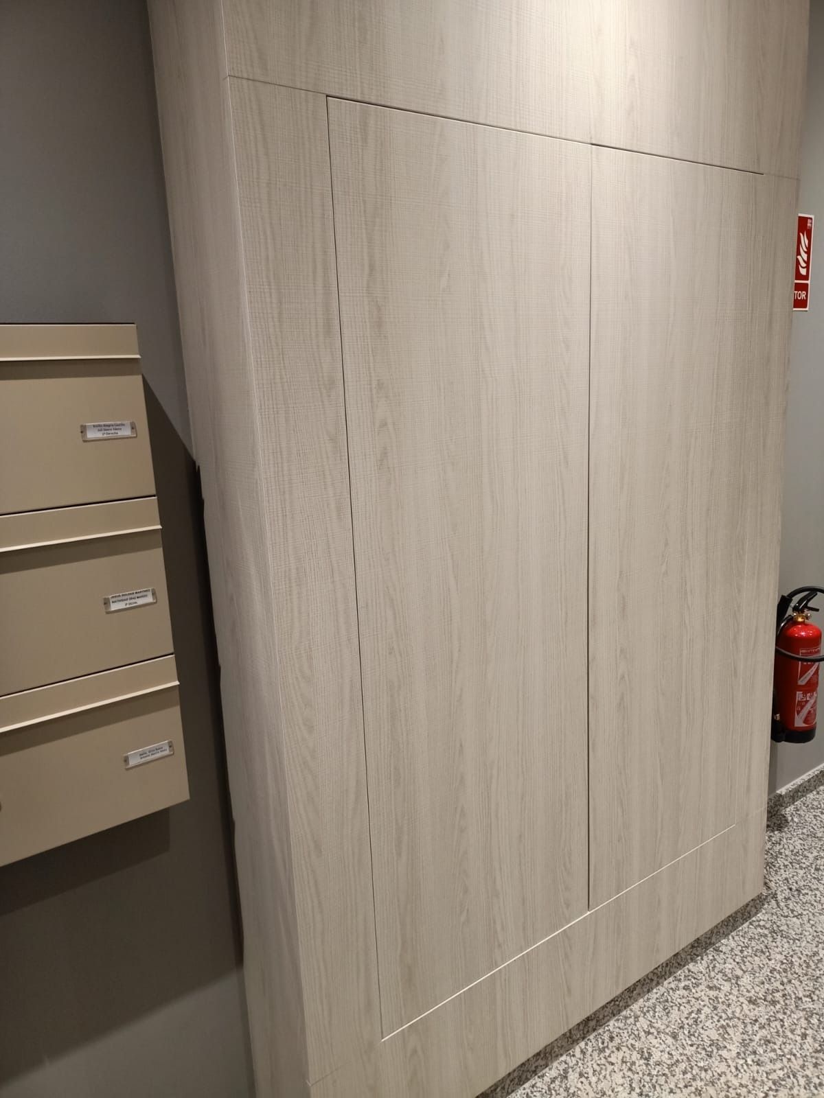En un pasillo, un gran armario de madera clara se encuentra junto a un conjunto de buzones metálicos y un extintor rojo.