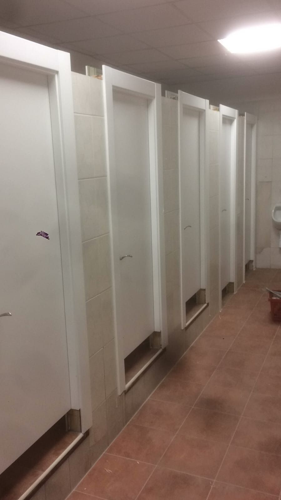 Una hilera de puertas blancas tipo cubículo, con aberturas en la parte inferior, ubicadas en un baño público con azulejos.