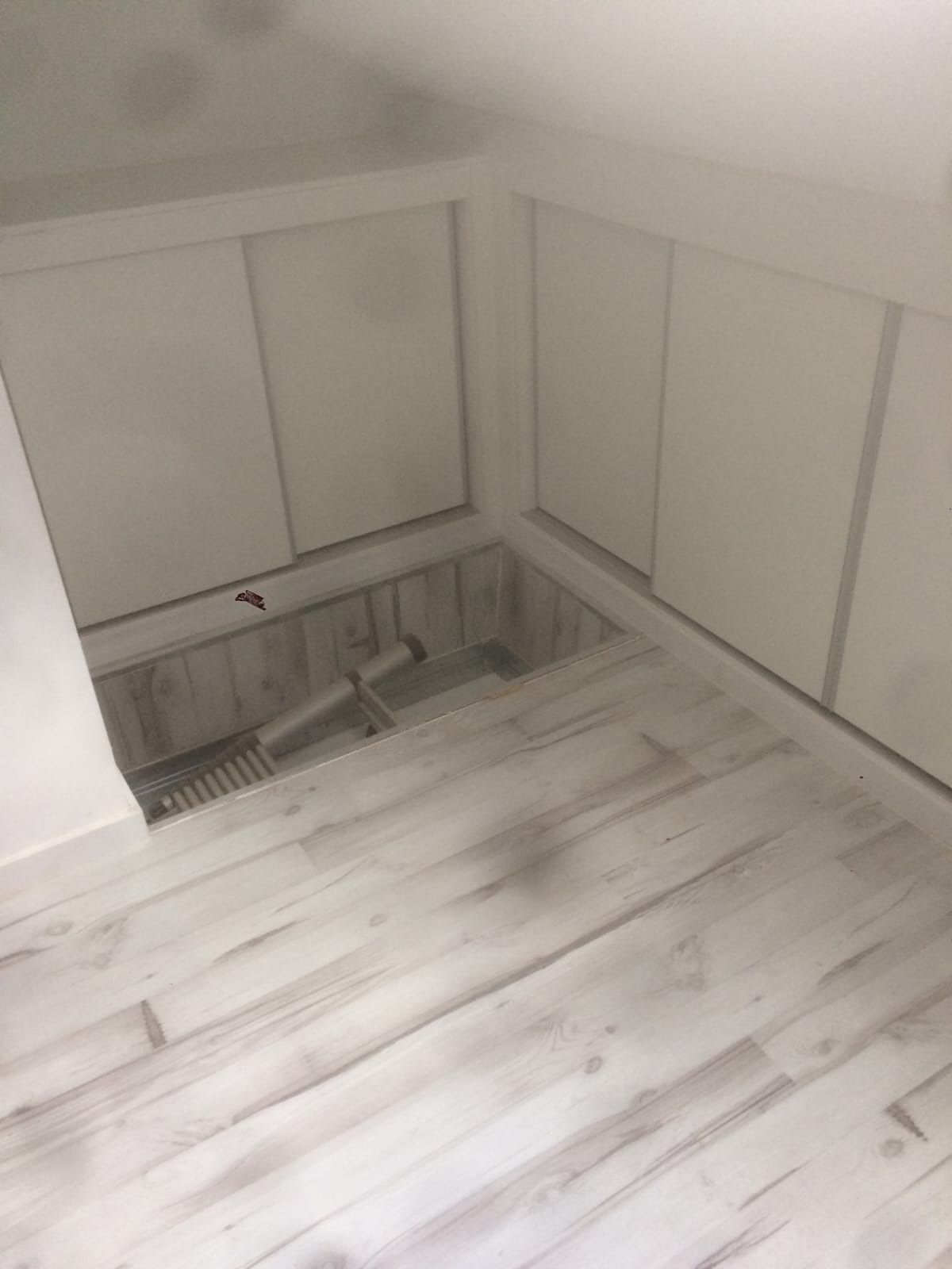 Una habitación con paredes blancas, suelo que imita la madera y un hueco en el suelo que contiene el mango de la aspiradora.