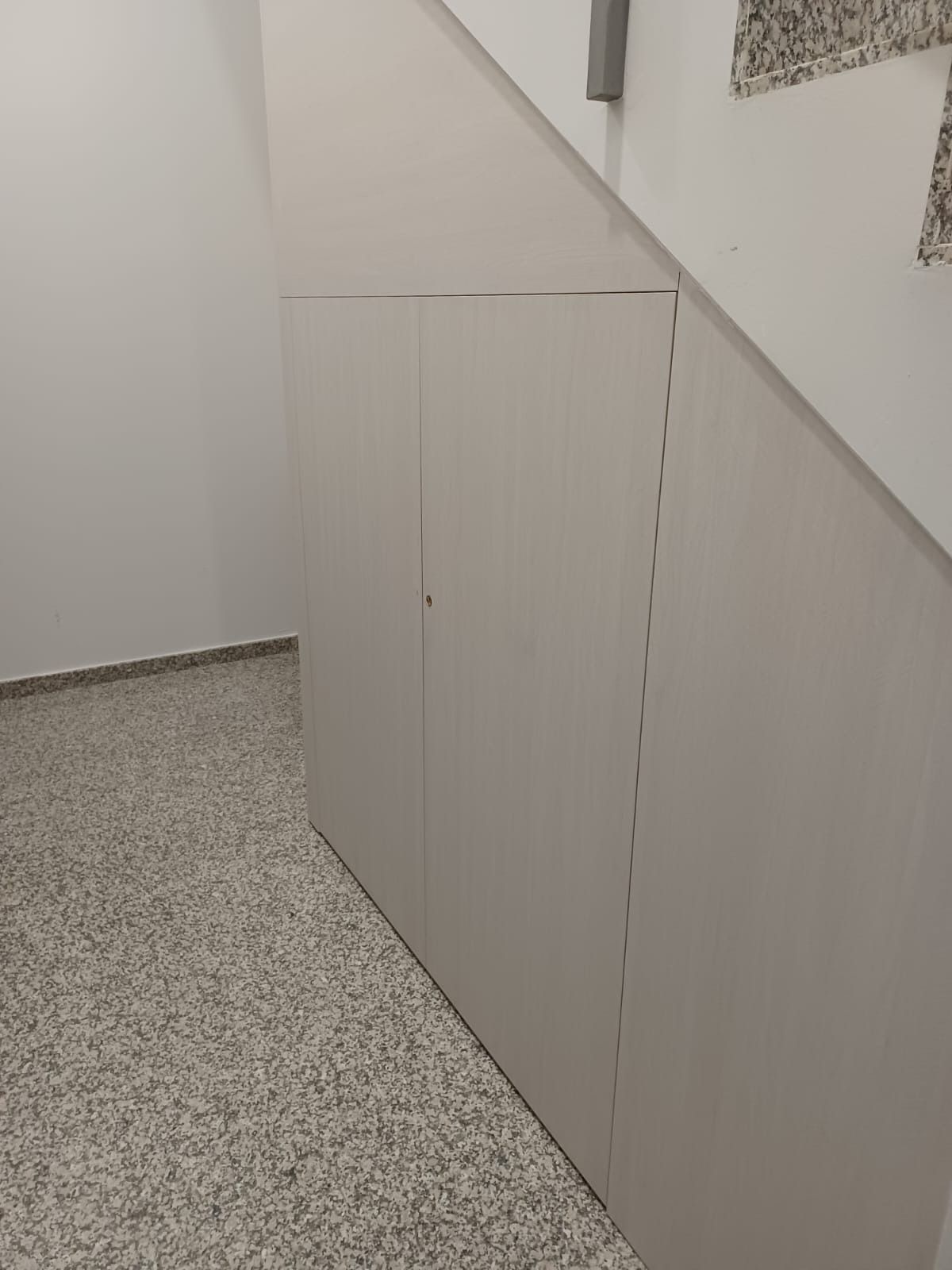 Un armario de almacenamiento blanco de dos puertas instalado debajo de una escalera en una habitación con suelo moteado.