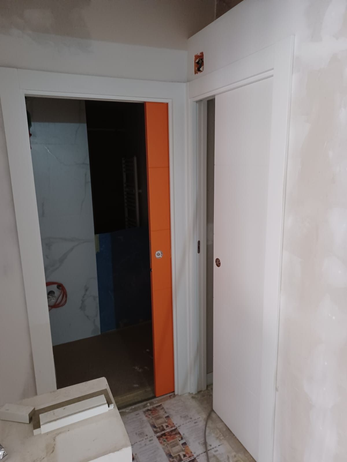 Un pasillo en obras que muestra una puerta abierta con un marco naranja junto a una puerta corredera blanca cerrada.