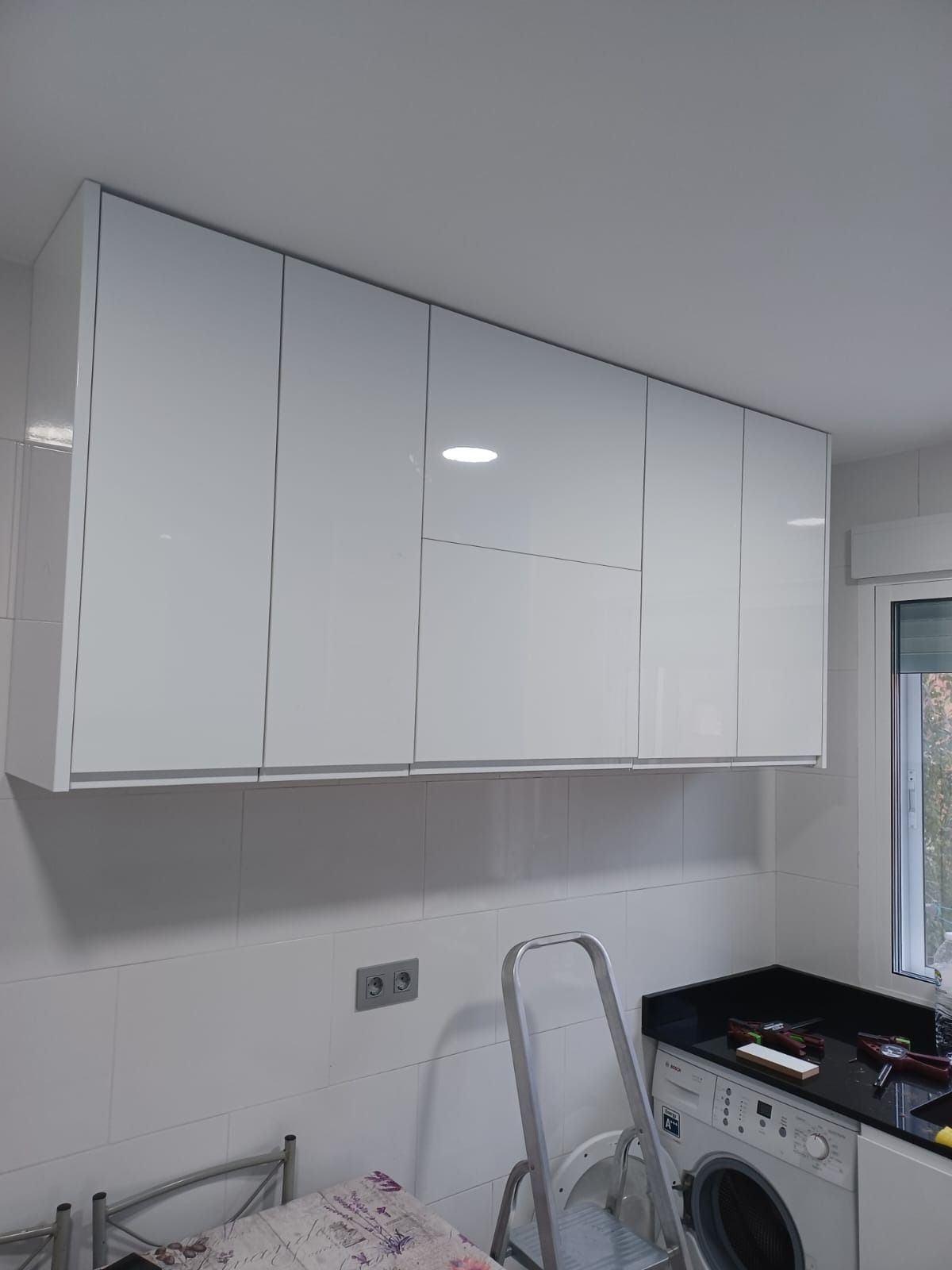 Armarios de cocina blancos montados sobre una pared con una toma de corriente visible, junto a una lavadora y una escalera.