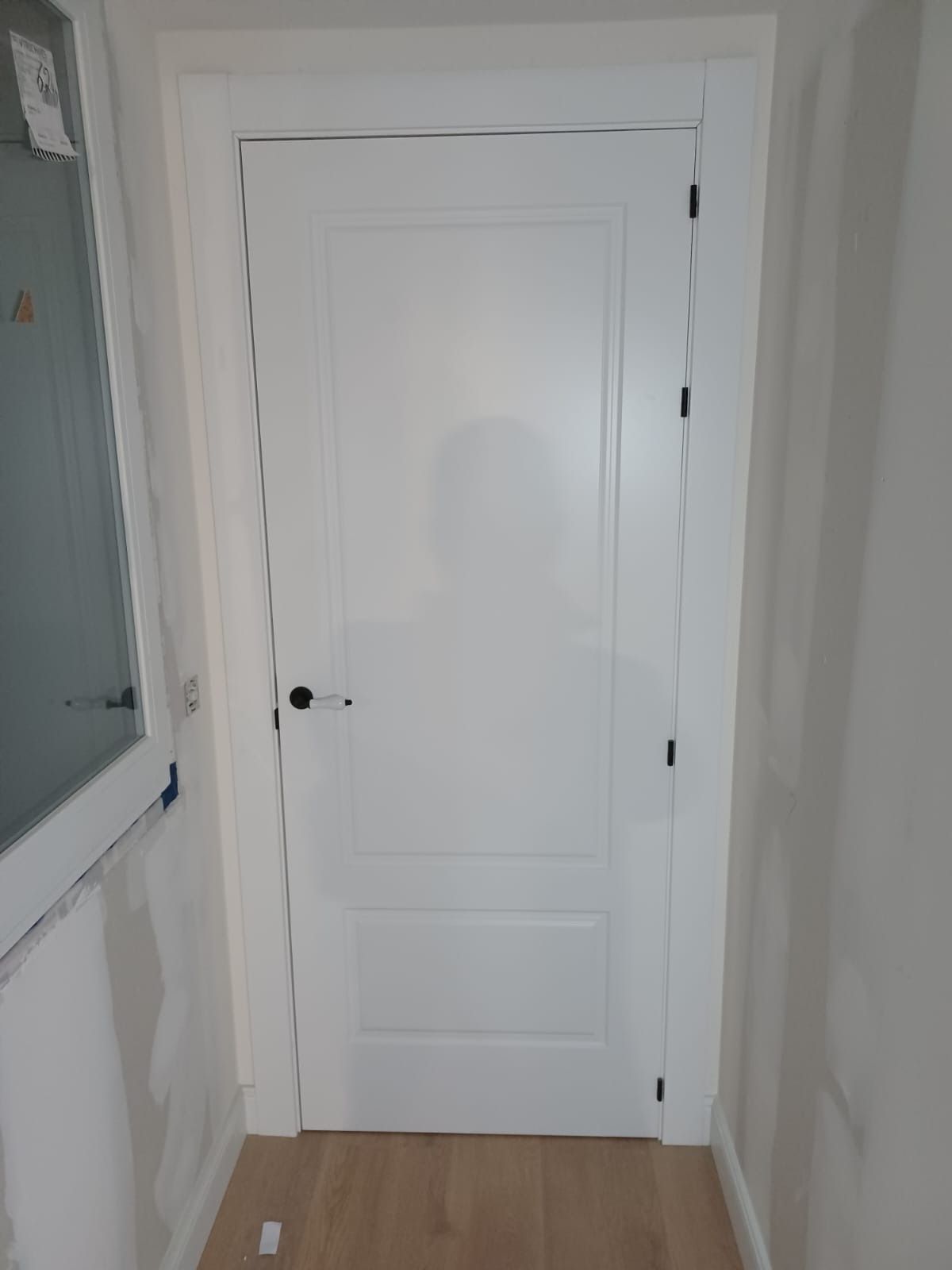 Una puerta interior blanca con paneles y tirador negro, situada en un pasillo de color claro con paredes sin terminar.