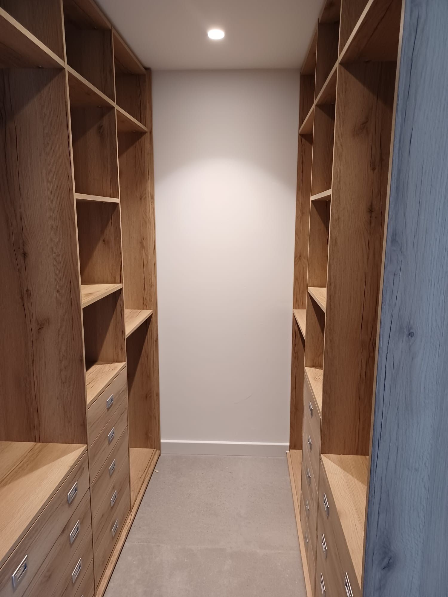 Un vestidor con estantes y cajones de madera clara a ambos lados, y una pared trasera blanca con una lámpara de techo.