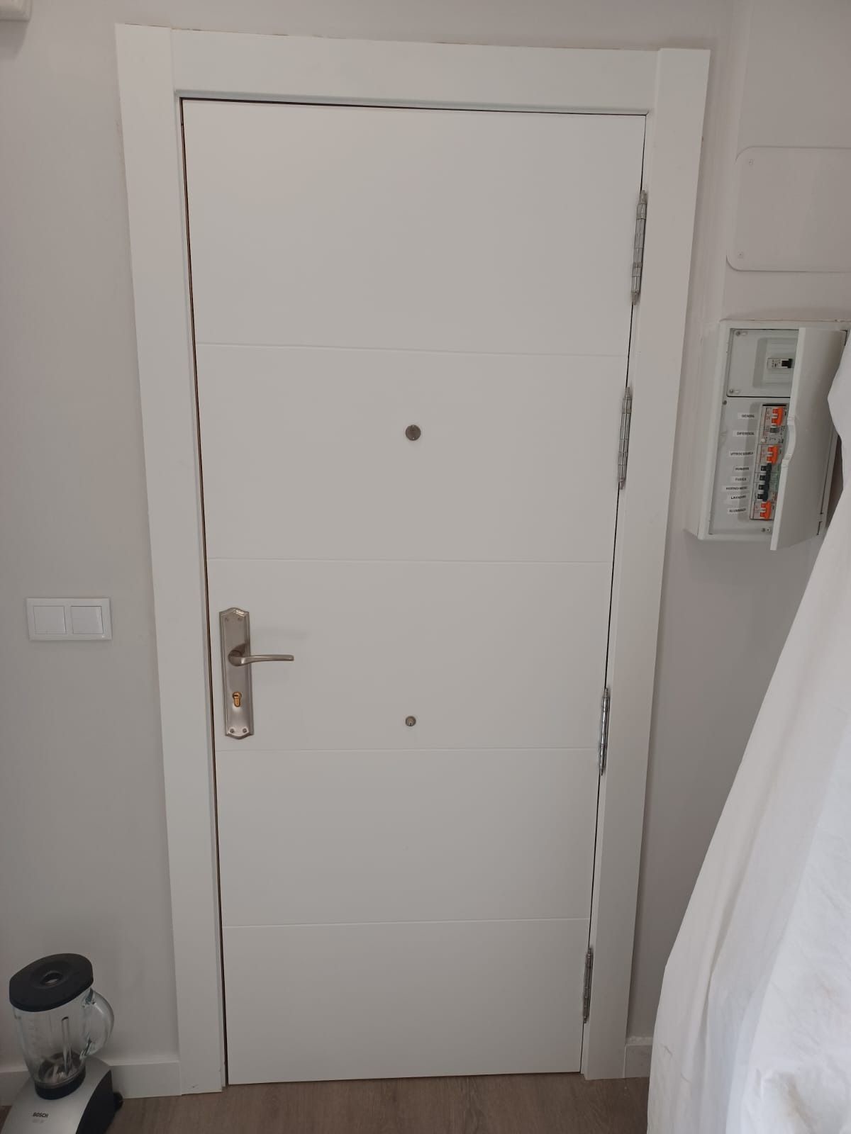 Una puerta interior blanca con manija plateada, dos mirillas y un panel eléctrico visible en la pared contigua.