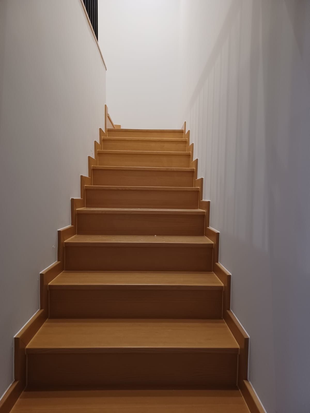 Vista frontal de una escalera de madera que asciende entre dos paredes blancas lisas.