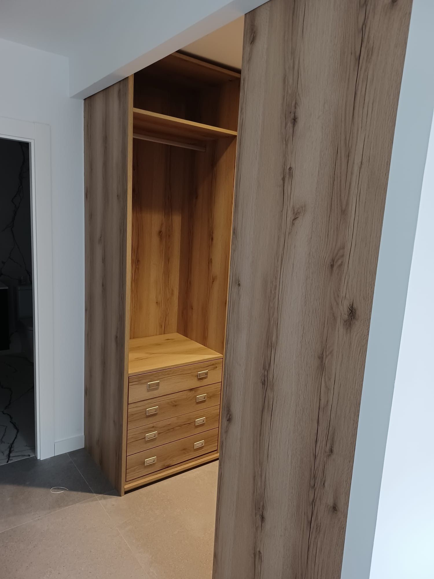 Un armario de madera abierto con barra para colgar ropa, estante superior y tres cajones, visto a través de una puerta.