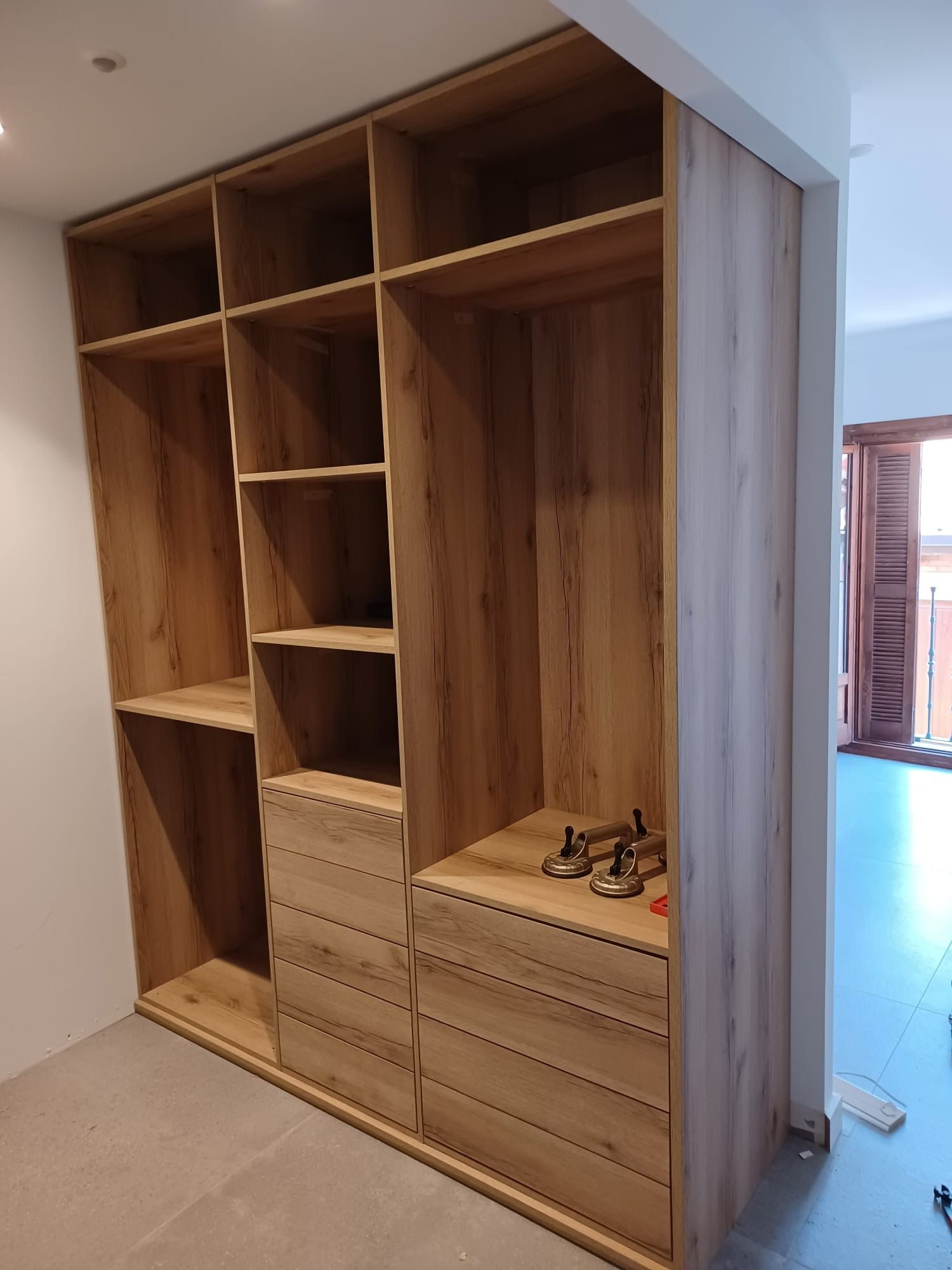 Un armario alto de madera clara con varios estantes abiertos y dos cajoneras, instalado en una habitación.