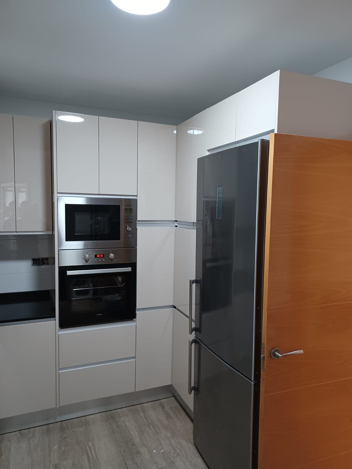 Una cocina moderna con armarios blancos, microondas y horno empotrados, frigorífico de acero inoxidable y puerta de madera.