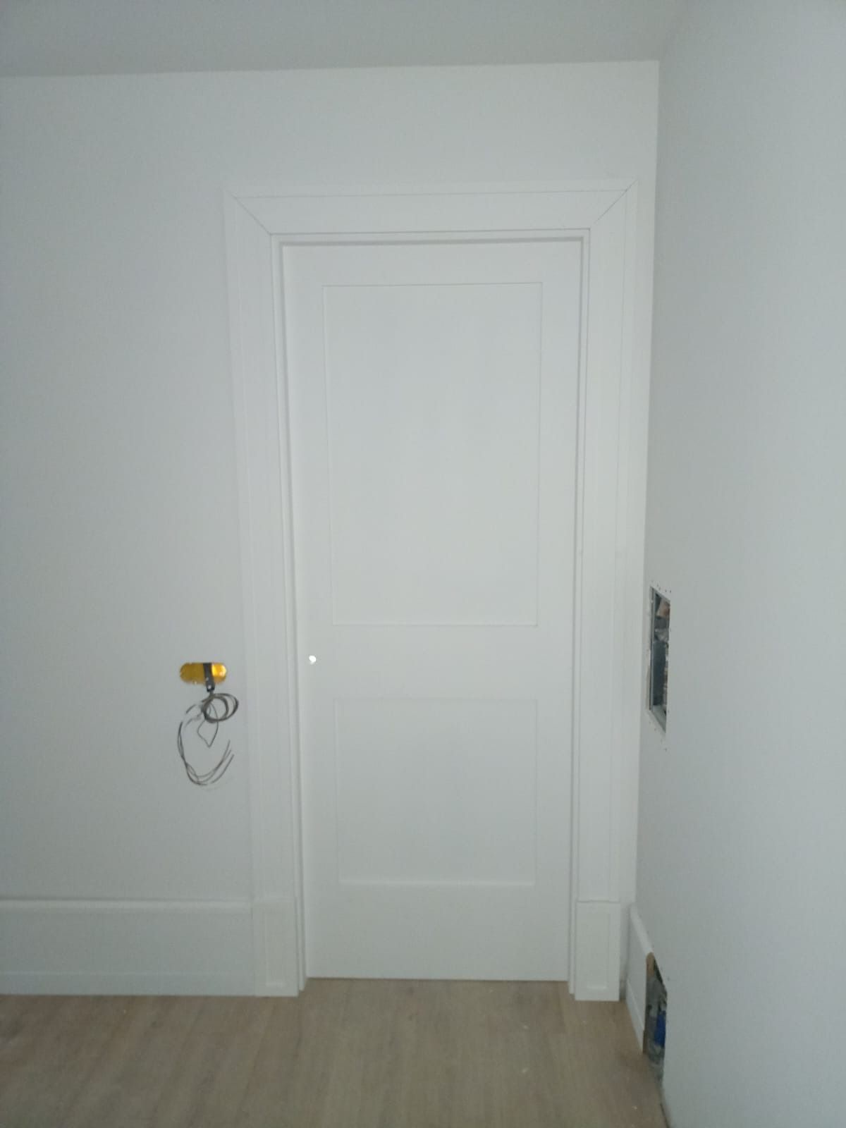 Una puerta interior blanca con herrajes y molduras de latón en una habitación con suelo de color claro y paredes blancas.