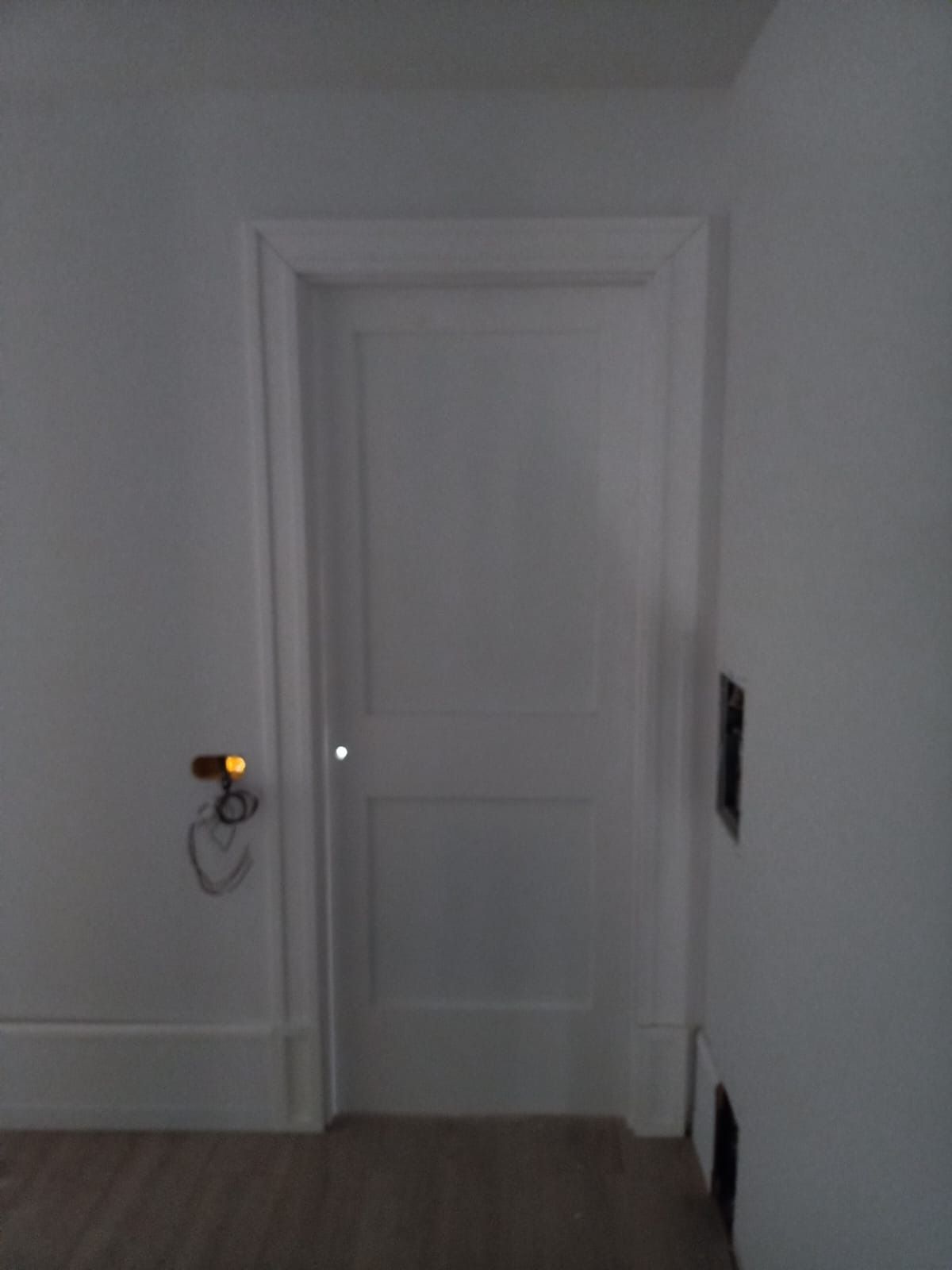 Una puerta interior blanca con marco blanco centrada en una pared blanca, con una pequeña lámpara a la izquierda.