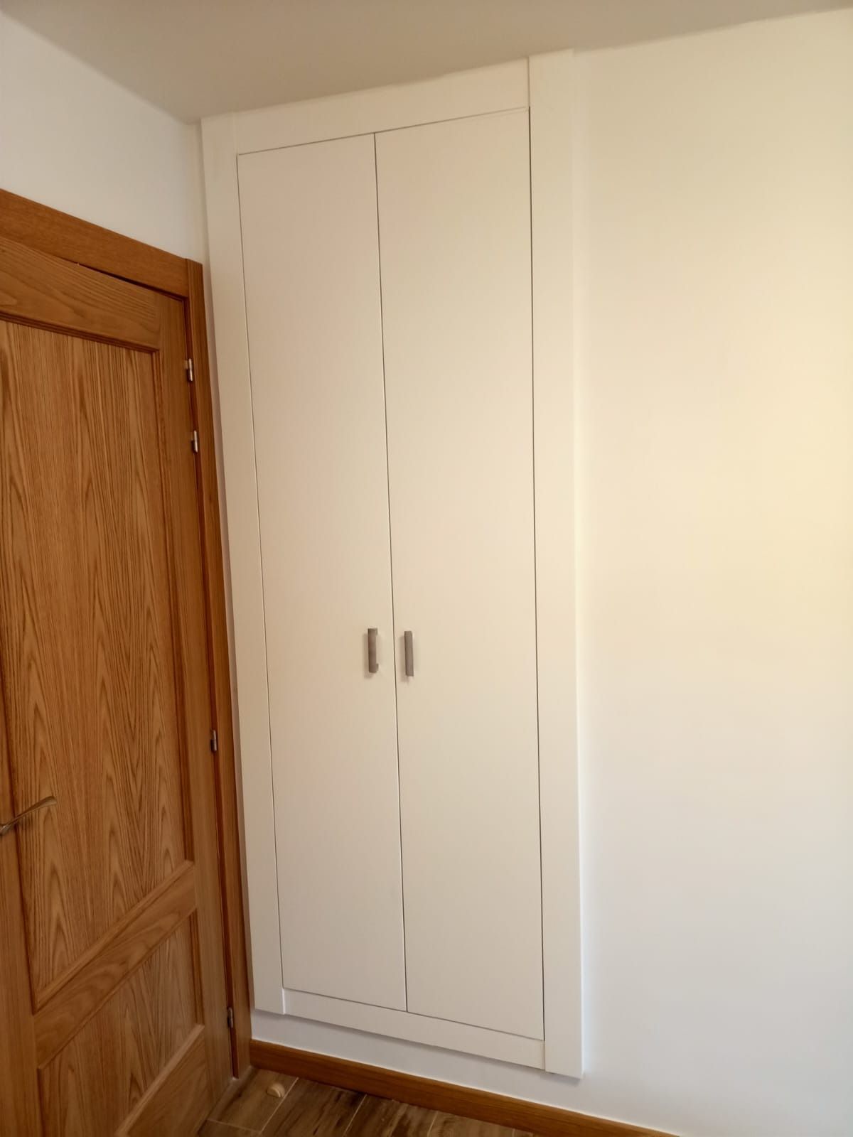 Un armario empotrado blanco con dos puertas y tiradores metálicos, situado junto a una puerta de madera en la pared.