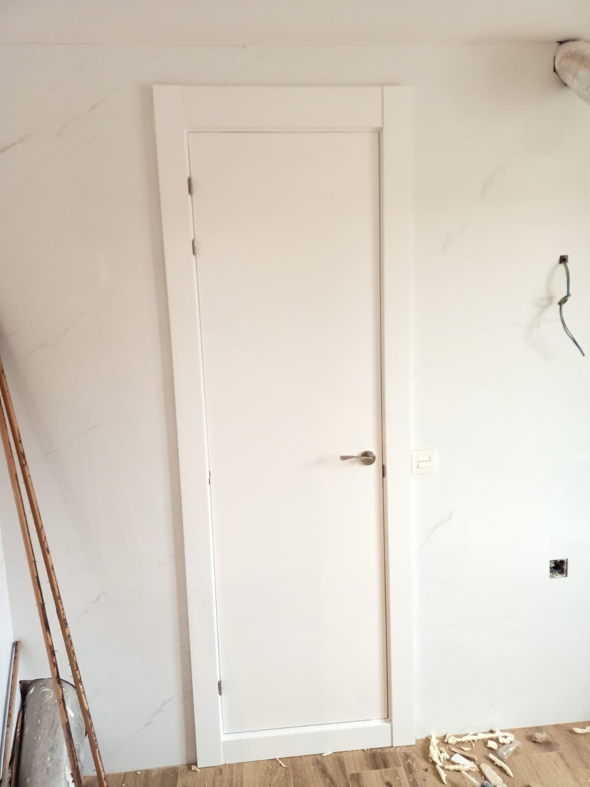 Una puerta interior blanca con manija plateada instalada en una habitación con paredes blancas y piso sin terminar.