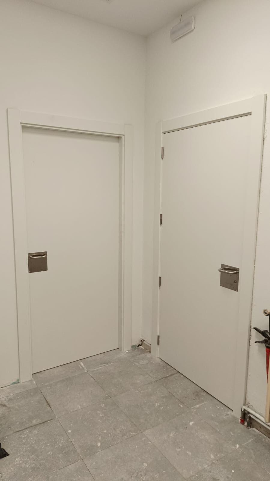 Dos puertas blancas con tiradores metálicos, una al lado de la otra, en una habitación con suelo de baldosas grises.