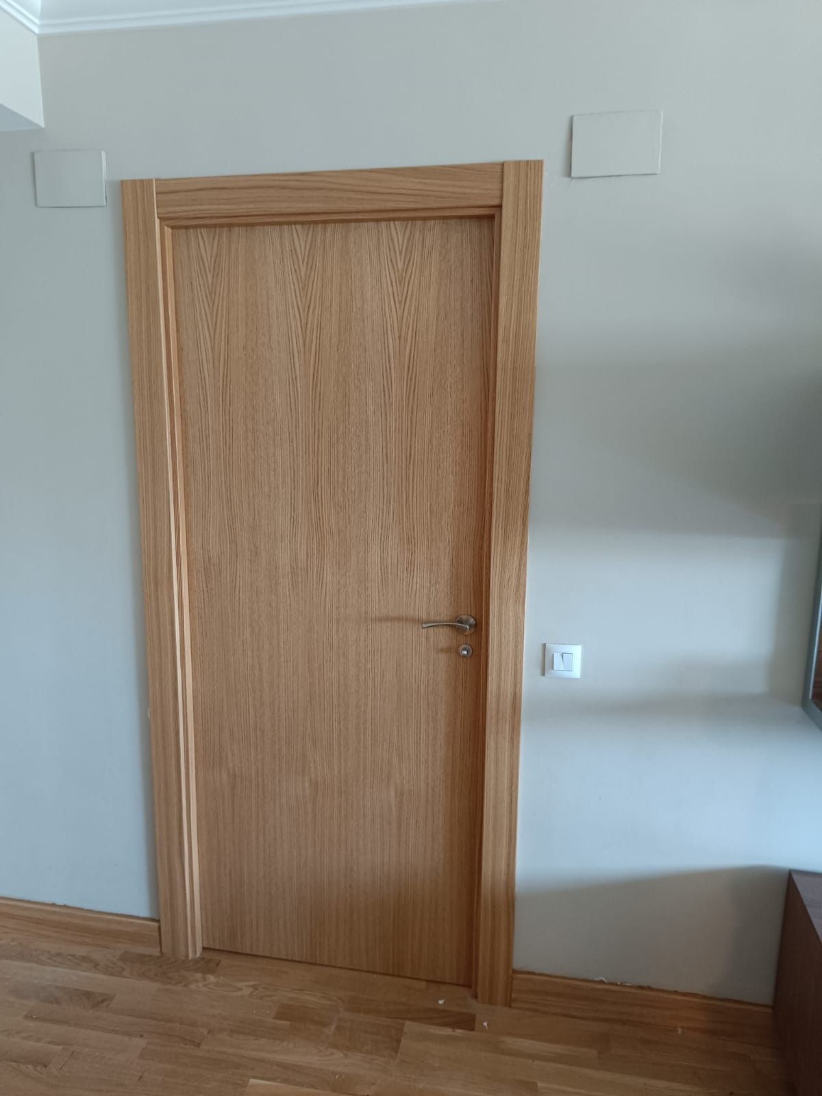 Una puerta de madera con molduras de tono claro y tirador plateado, situada en una pared gris claro sobre un suelo de madera.
