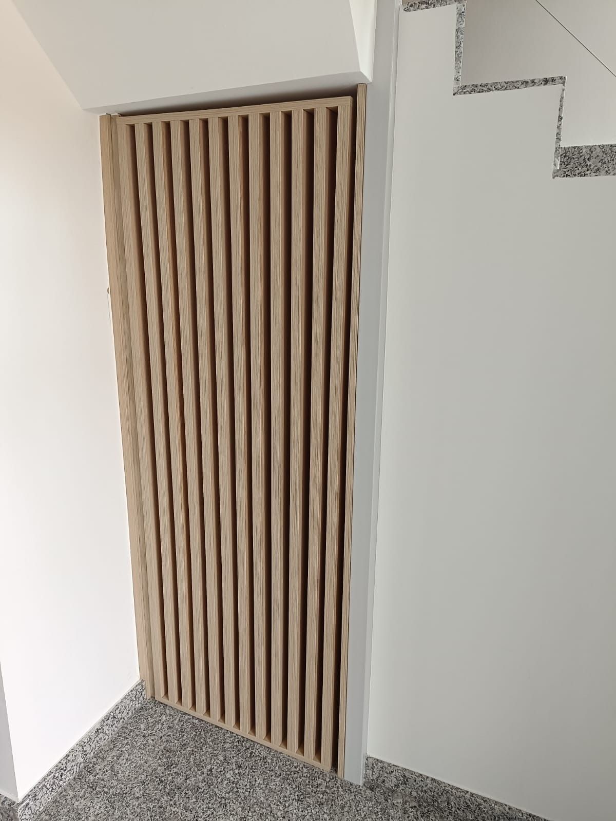 Una puerta de listones de madera clara instalada debajo de una escalera, junto a una pared blanca y un suelo gris moteado.