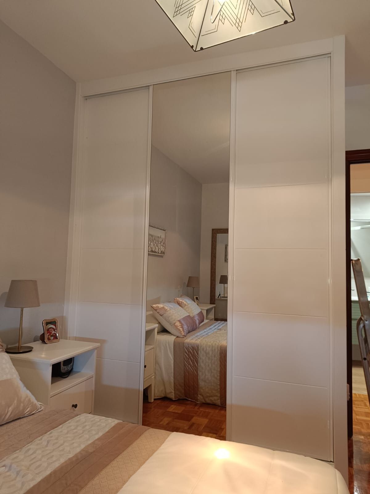 Un dormitorio con un gran armario blanco con un panel central espejado que refleja una cama y una puerta.