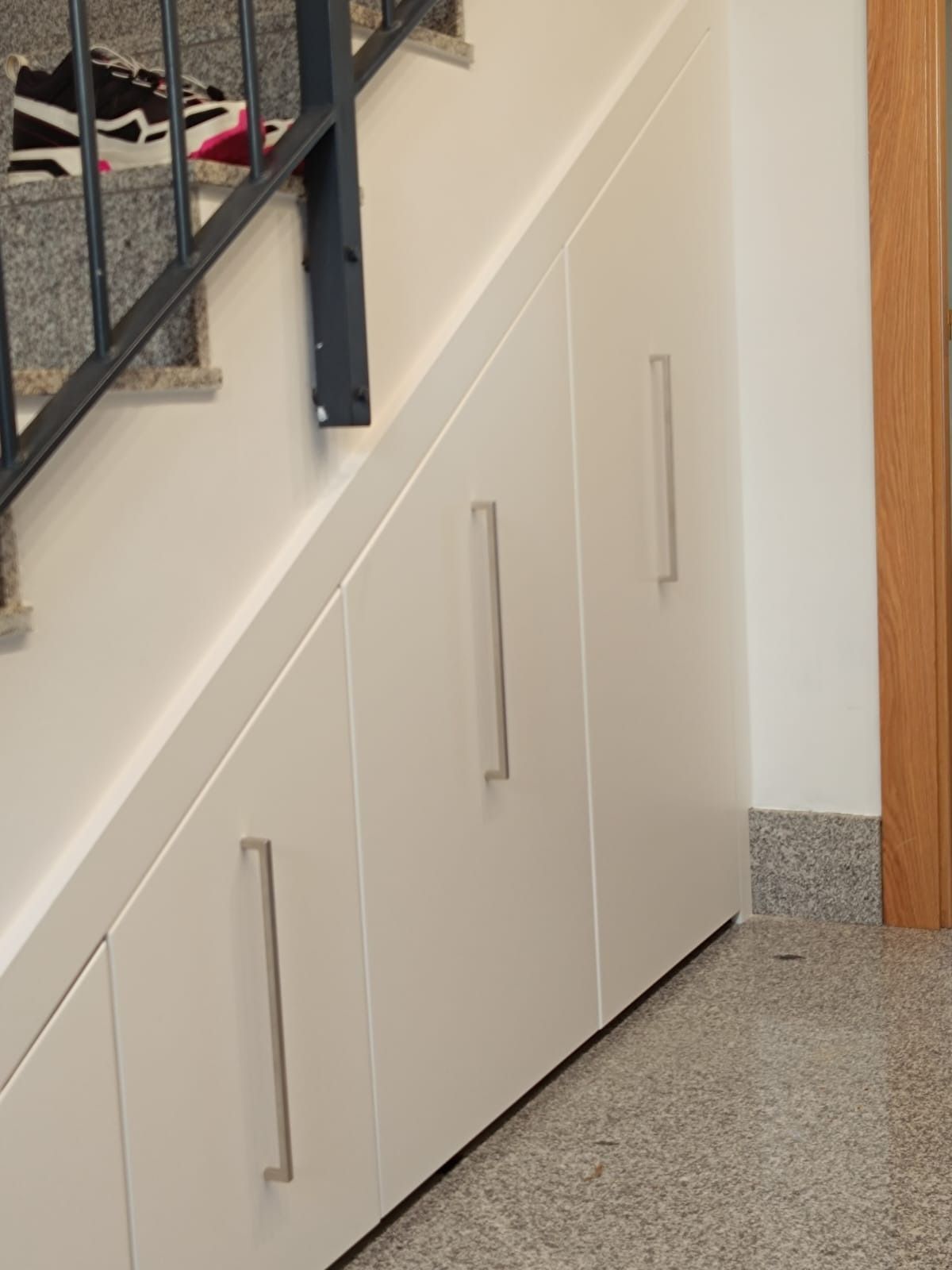 Armarios de almacenamiento blancos, hechos a medida, con tiradores verticales plateados, situados debajo de una escalera.