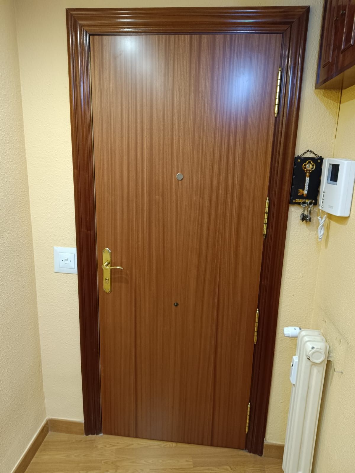 Una puerta interior de madera de color marrón con herrajes dorados, una mirilla y un radiador de color claro a su lado.