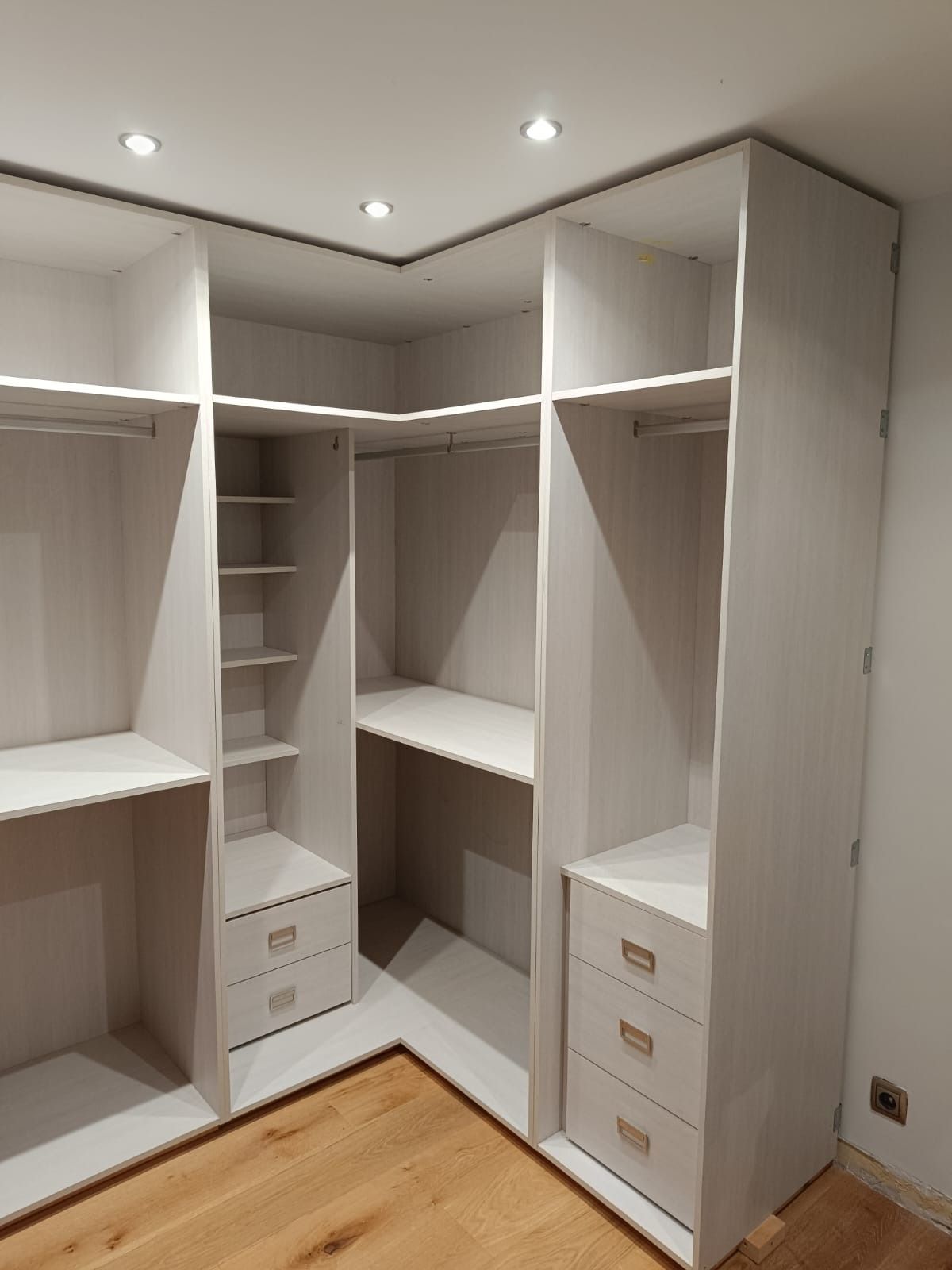 Un armario empotrado en forma de L, de madera clara, con estantes abiertos, barras para colgar ropa y cajones apilados.