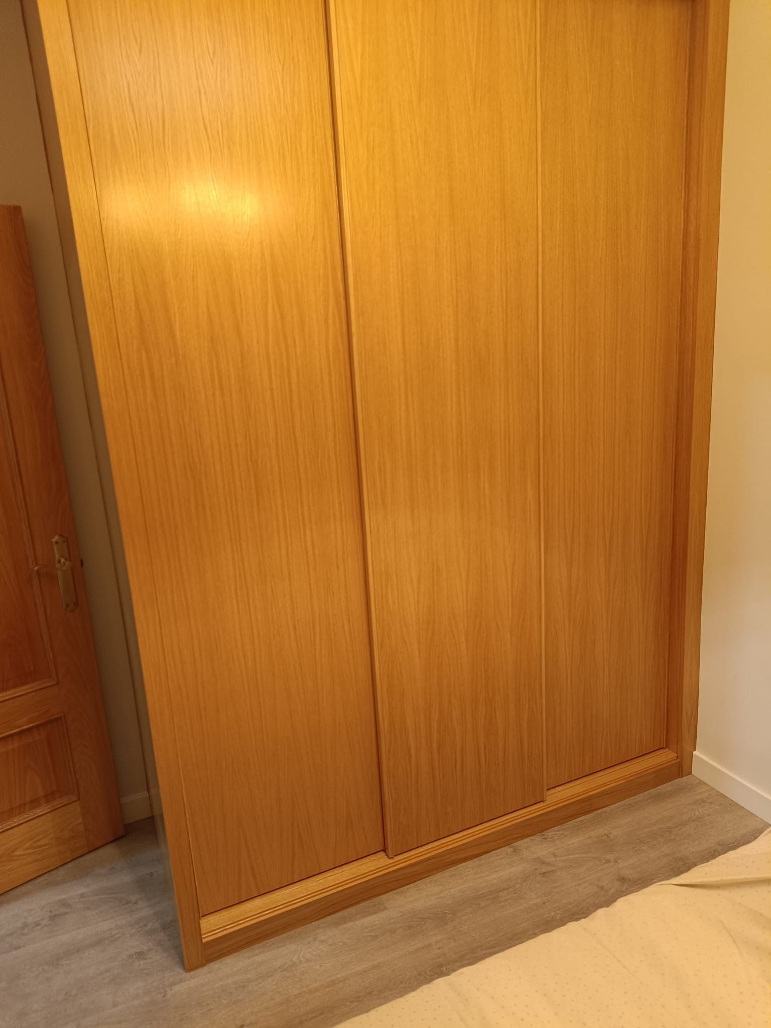 En una habitación con suelo de imitación madera de color gris claro, hay un armario de madera con tres paneles correderos.