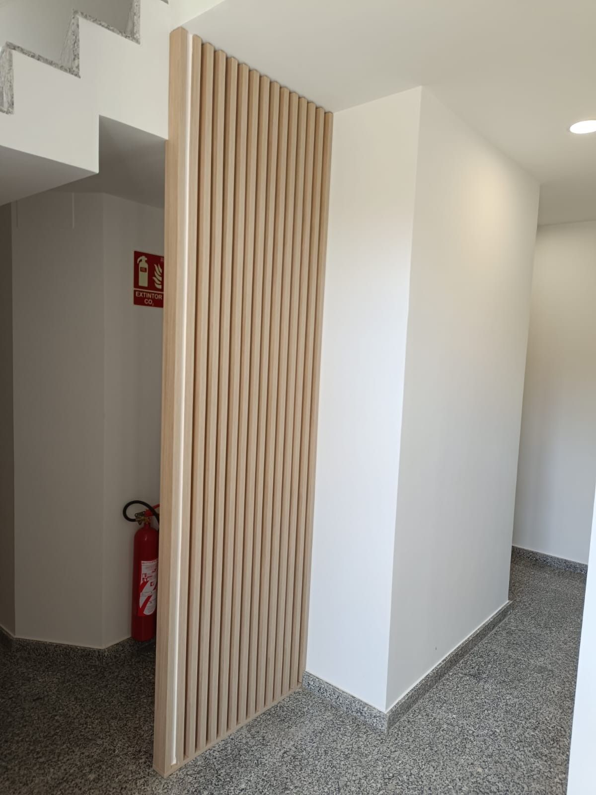 Una mampara de listones de madera clara separa un pasillo con suelo de granito gris y un extintor rojo de una escalera.