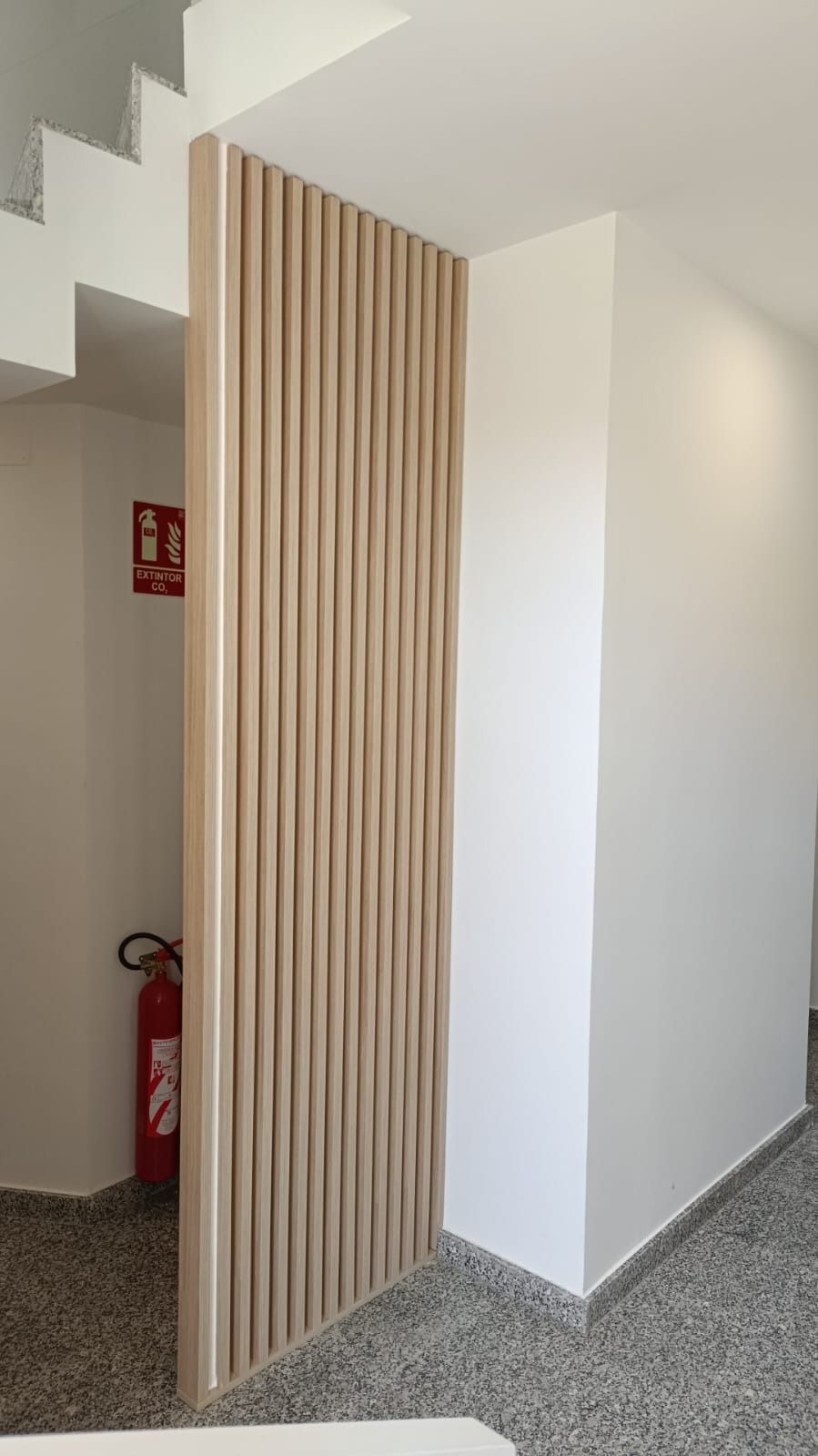 Un biombo de listones de madera se encuentra en un pasillo blanco, junto a una escalera y un extintor rojo.