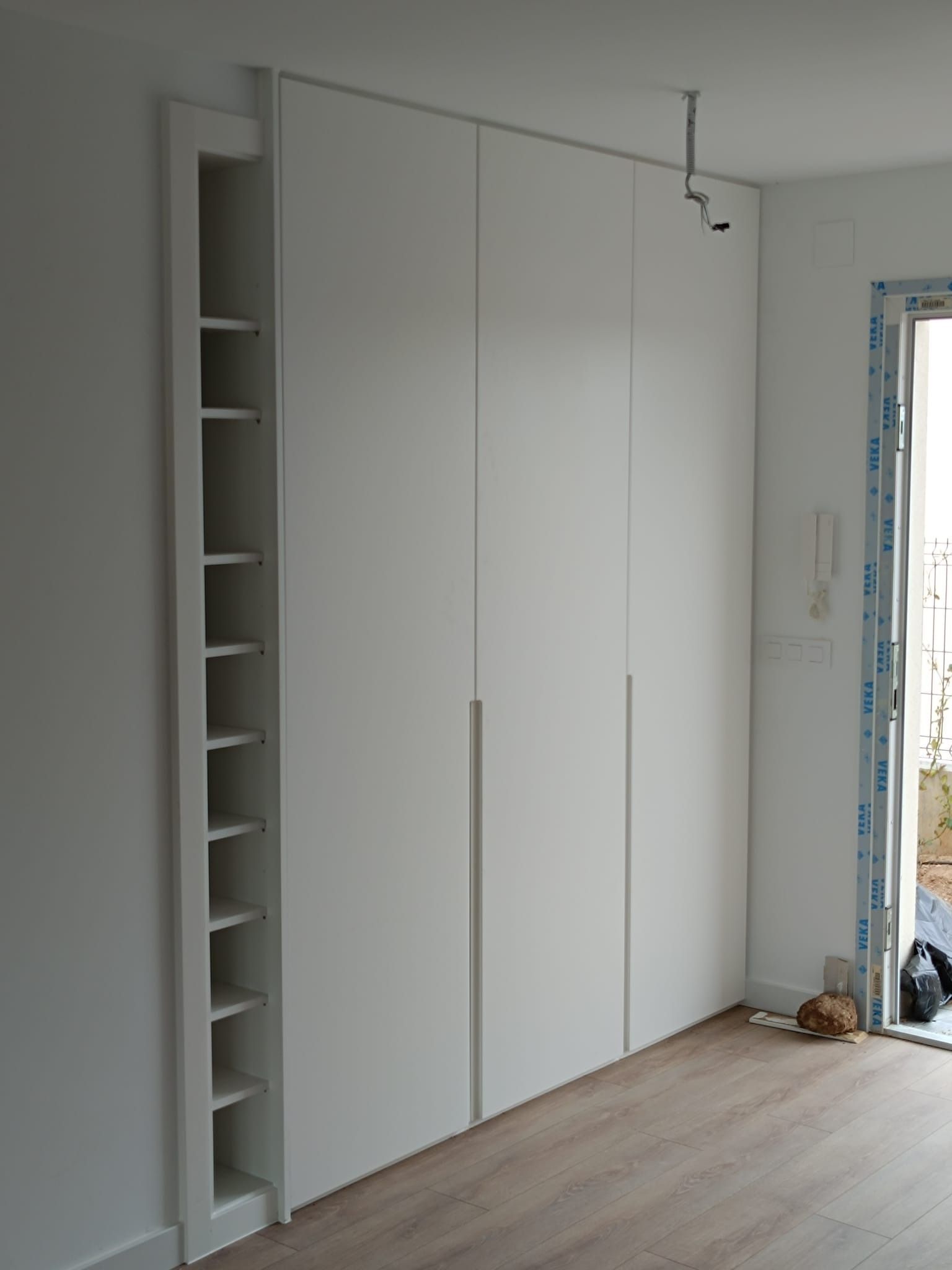 Un armario blanco que ocupa toda la pared, con tres puertas y una estantería vertical abierta integrada en el lado izquierdo.