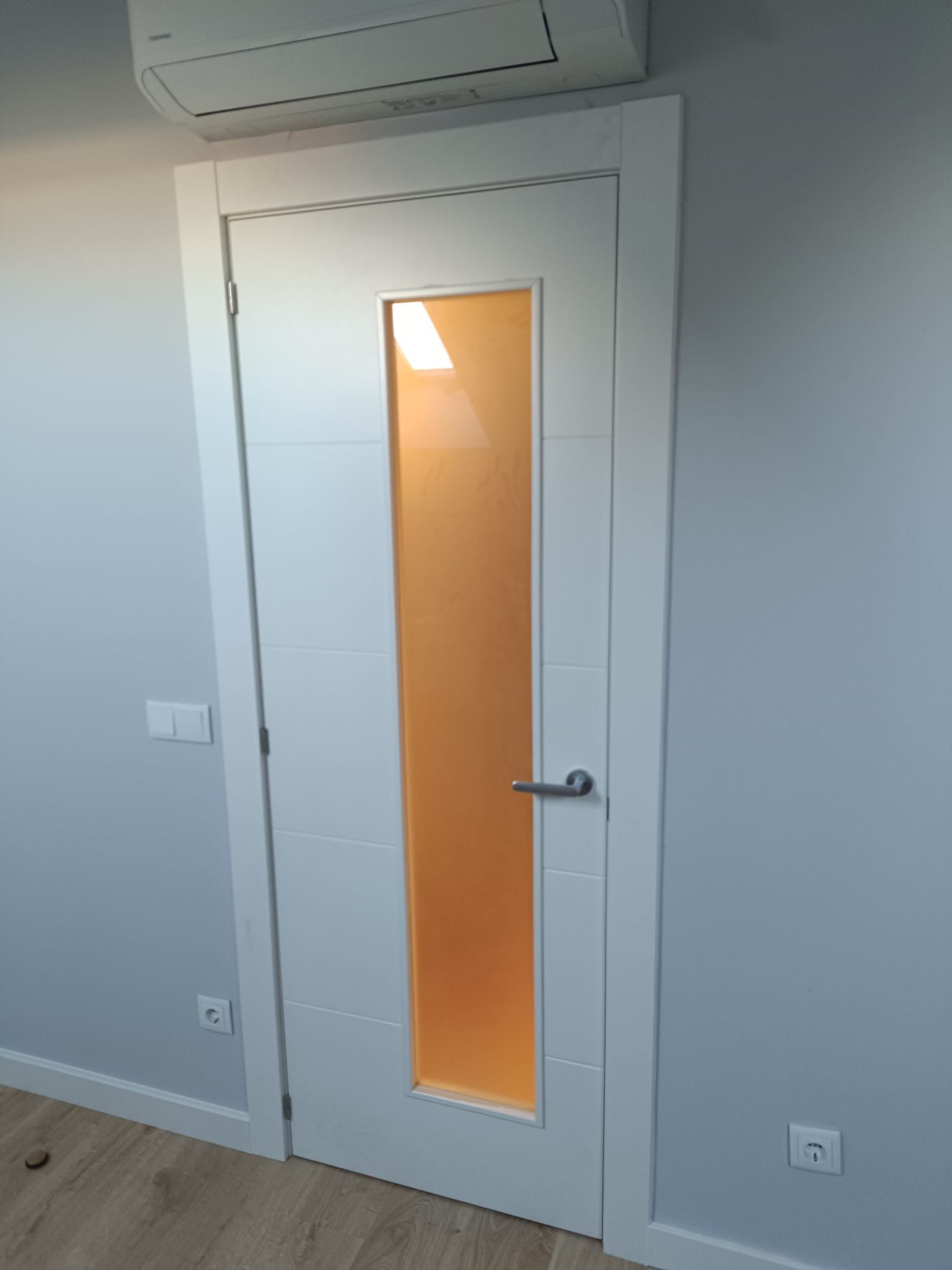 Una puerta interior blanca con un panel largo y vertical de vidrio esmerilado centrado en una pared gris claro.