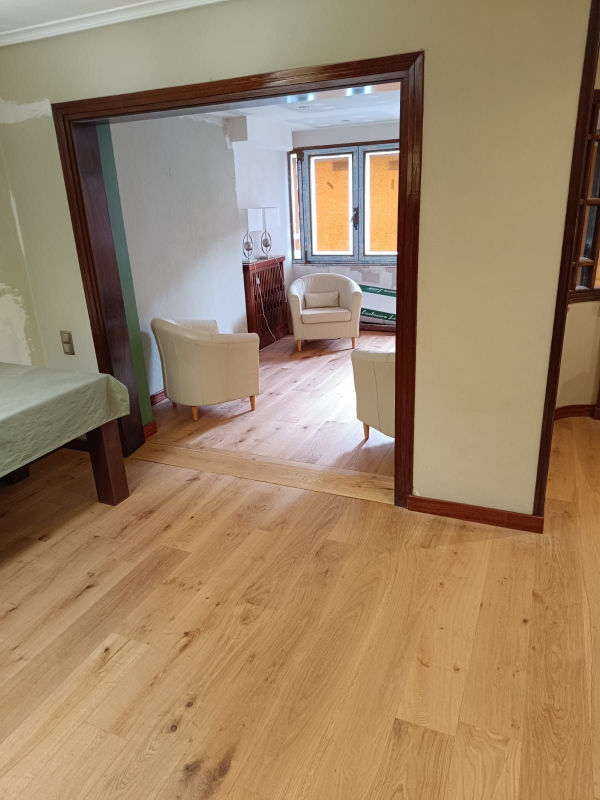 Vista interior a través de una puerta de madera oscura que da a una sala de estar con suelos de madera clara y tres sillones.