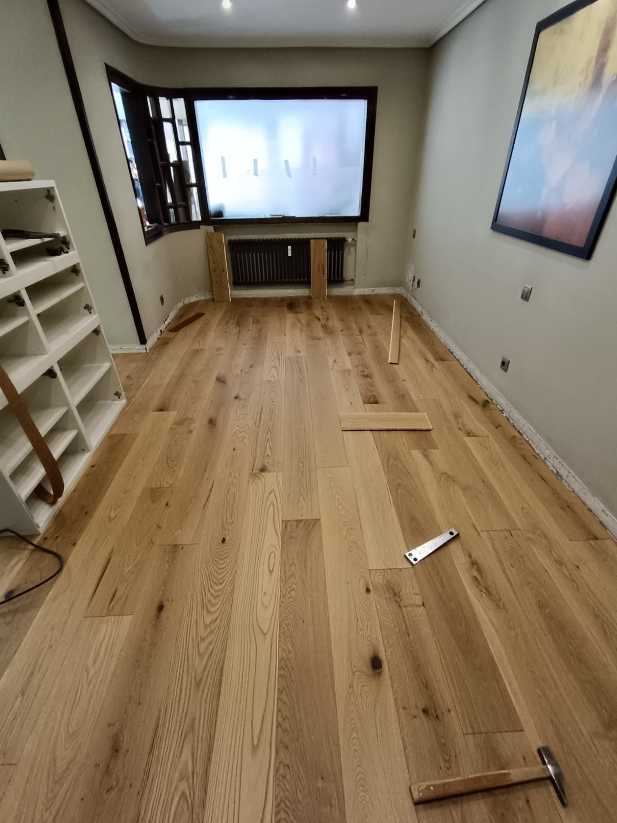Una habitación en obras con suelo de madera clara recién instalado, herramientas en el suelo y una estantería a la izquierda.