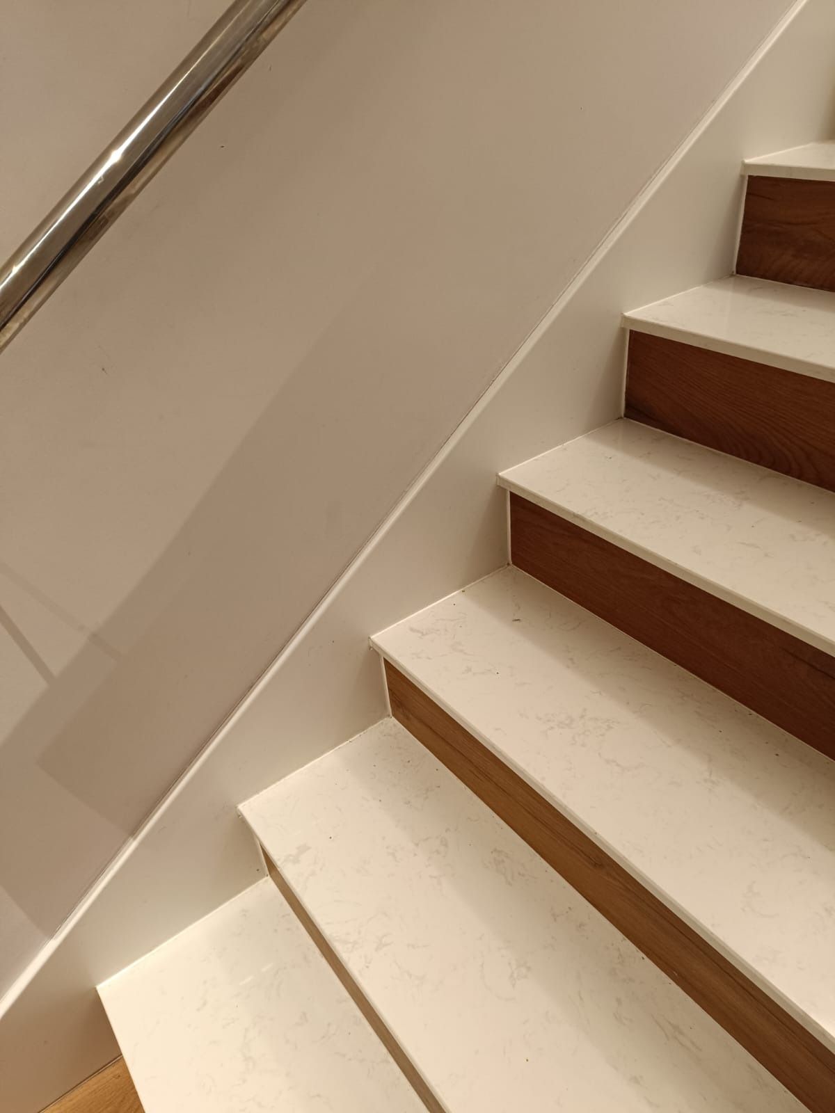 Una escalera con peldaños de mármol blanco, contrahuellas de madera y una barandilla metálica fijada a una pared blanca.