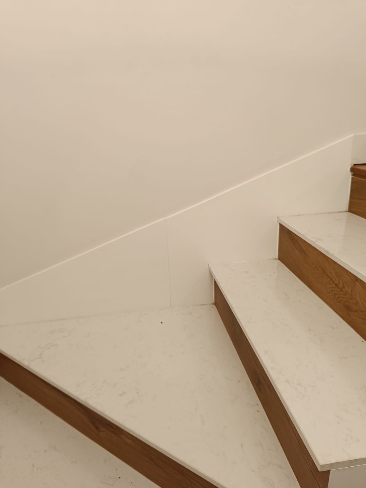 Escalera con peldaños de piedra blanca, contrahuellas de madera y un remate diagonal blanco a juego en la pared.