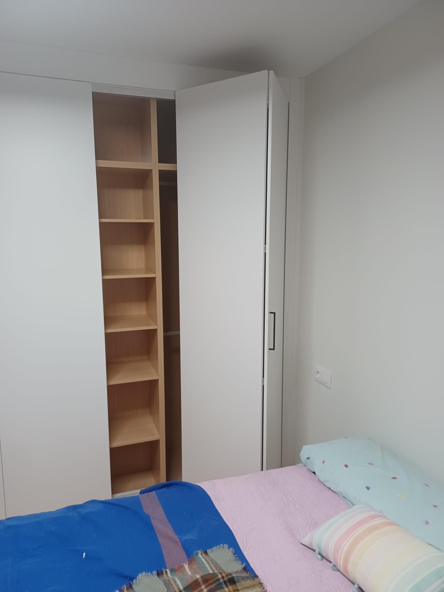 Un armario blanco con una sección abierta que deja ver estantes de madera, junto a una cama con ropa de cama azul y rosa.