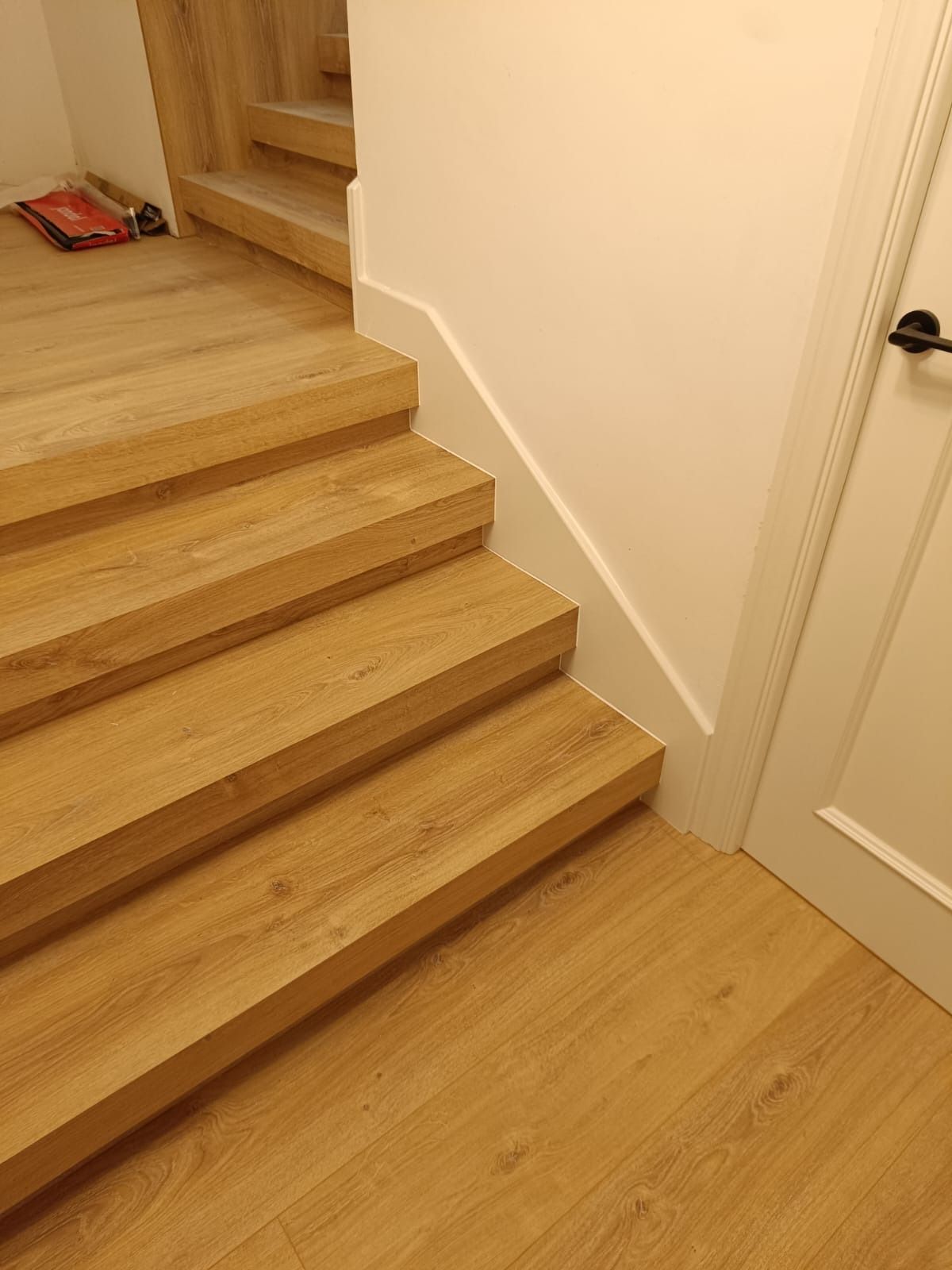 Una escalera de madera clara que asciende junto a una pared blanca y una puerta cerrada.