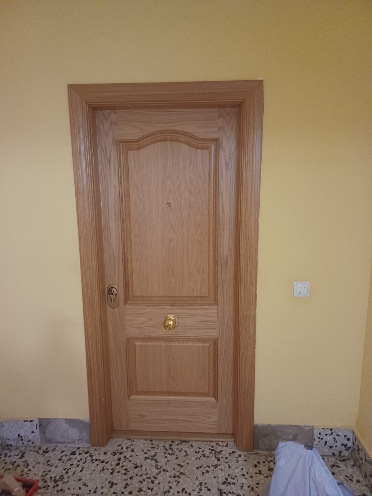 Una puerta de madera de color roble claro con paneles y herrajes dorados, centrada en una pared lisa de color amarillo claro.