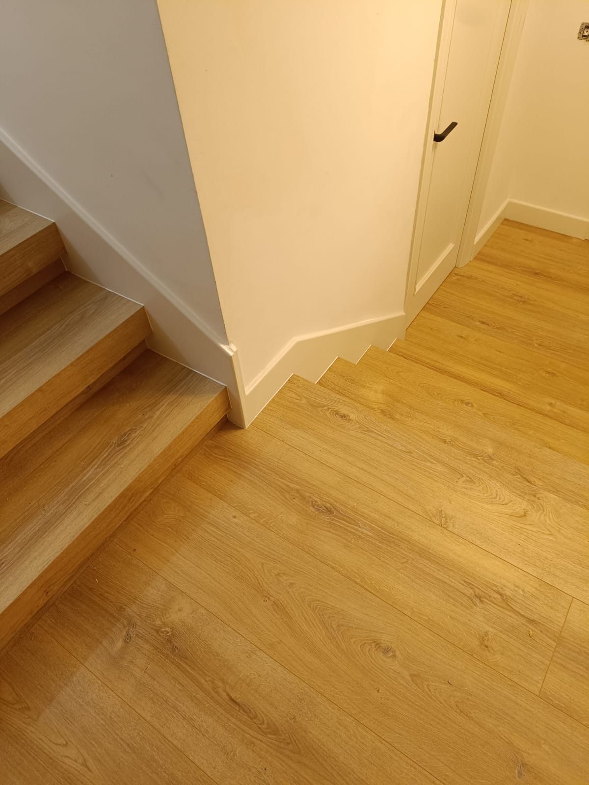 Escalera de madera clara que desciende hasta un suelo a juego, junto a una pared y una puerta blancas.