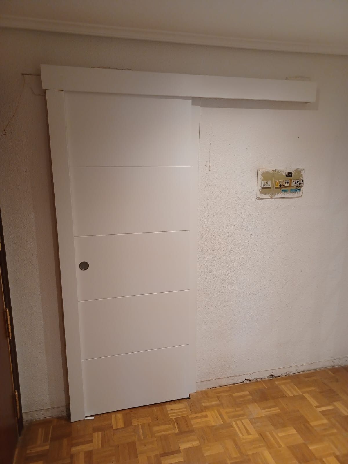 Una puerta corredera blanca con paneles horizontales, montada en una pared sobre un suelo de parqué de madera.
